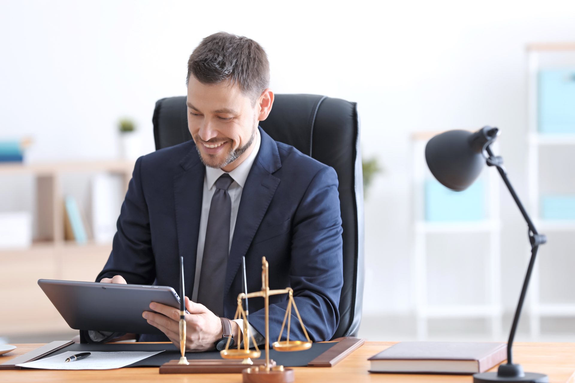 Person in blue suit sitting at a desk holding an ipad and smiling