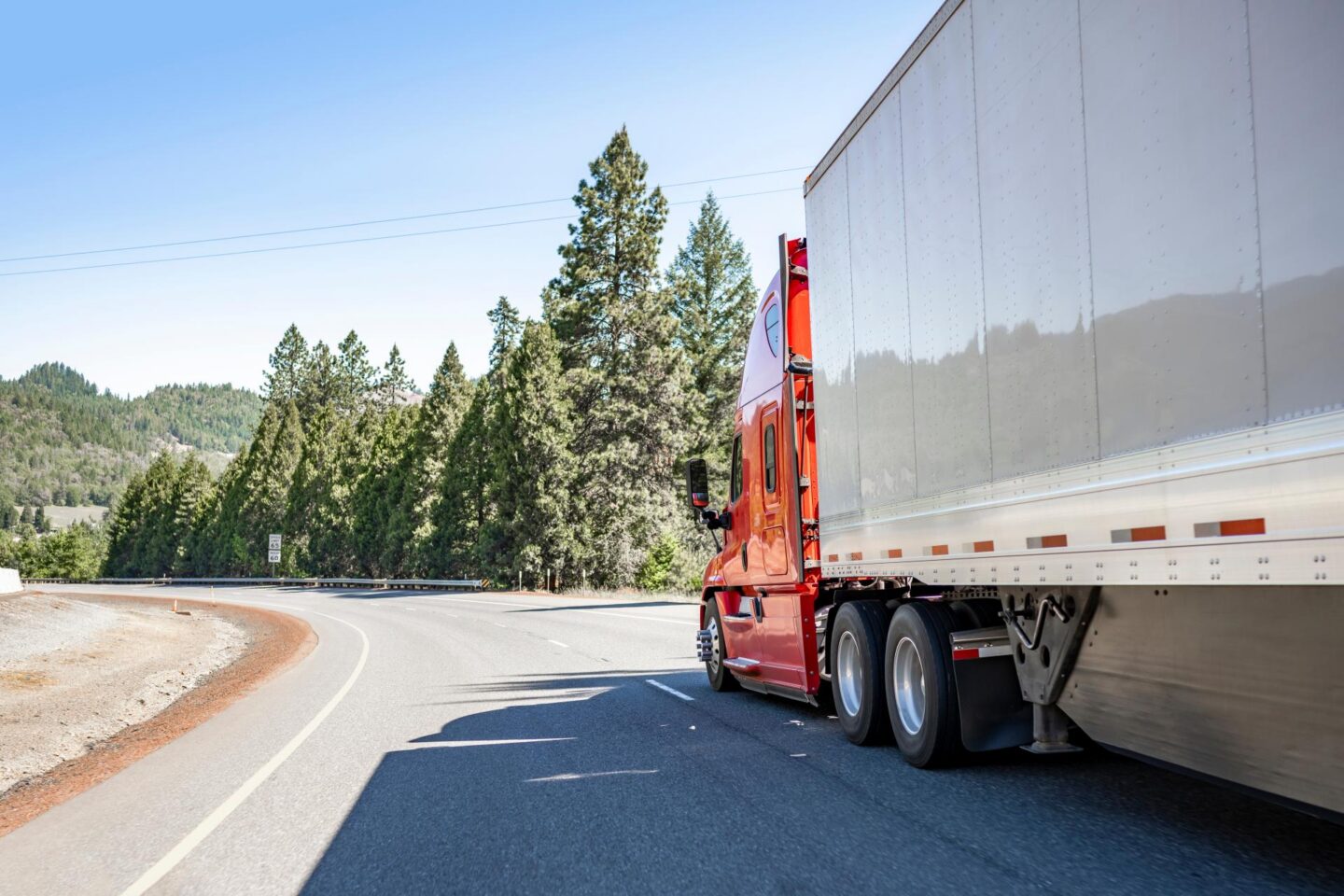 A red semi truck with a white trailer drives on a route near green trees.