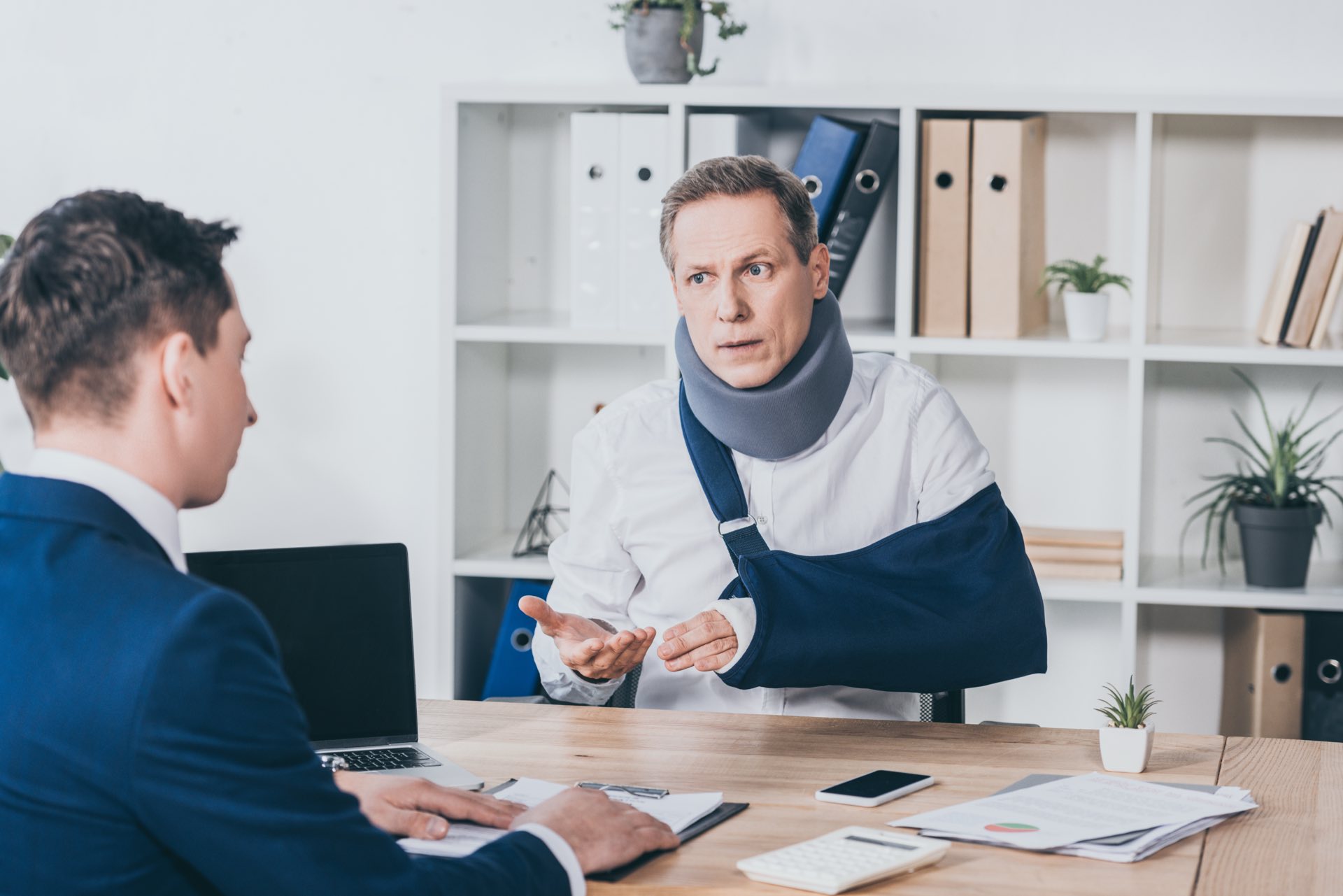 Person in neck brace with a broken arm witting at a desk talking with someone in a business suit with papers on the desk