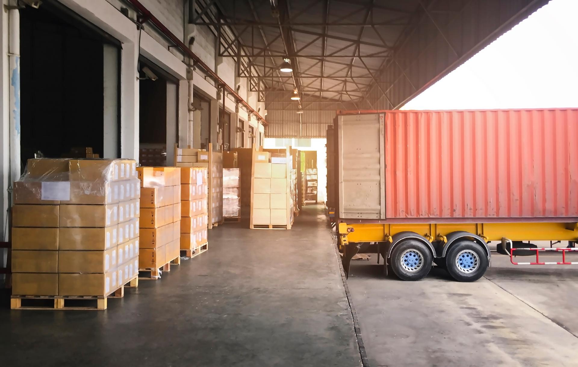 A cargo truck backed in to a loading bay with boxes stacked next to it.
