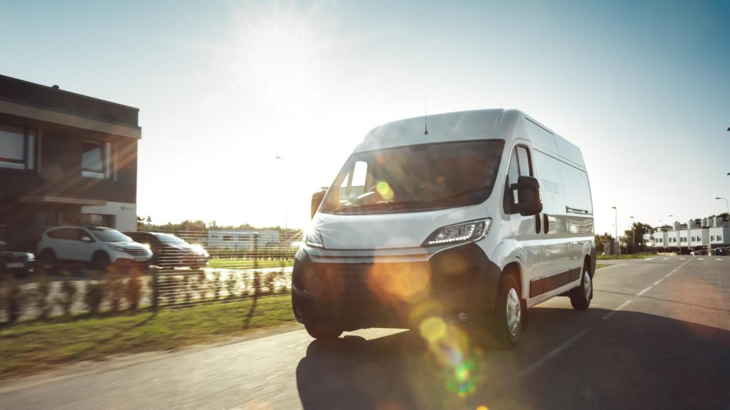 A white commercial van drives down a sunny road next to a business.