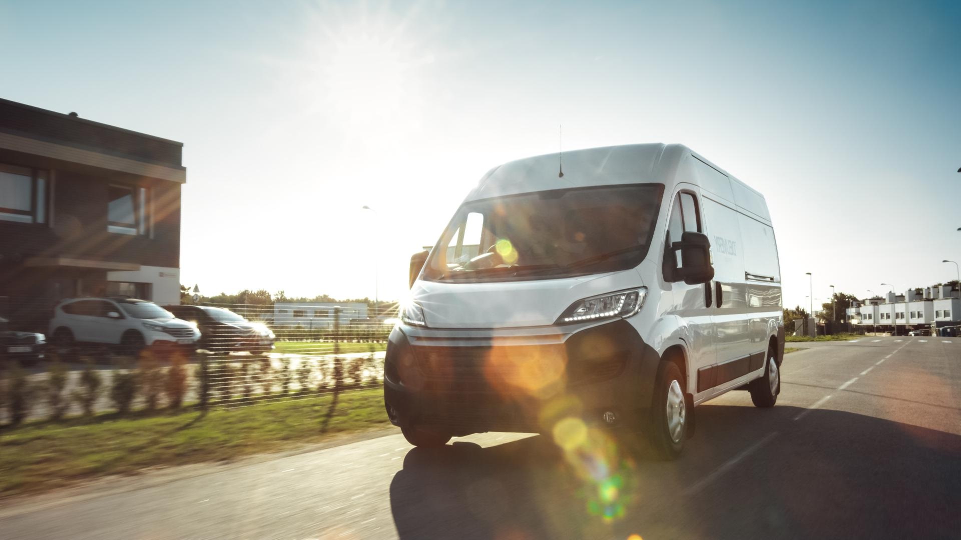 A white commercial van drives down a sunny road next to a business.