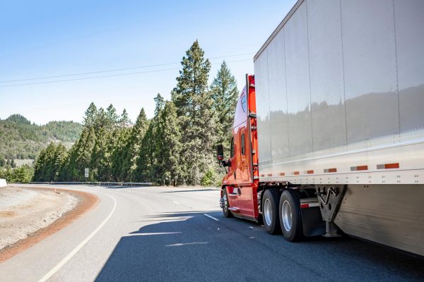 A red semi truck with a white trailer drives on a route near green trees.