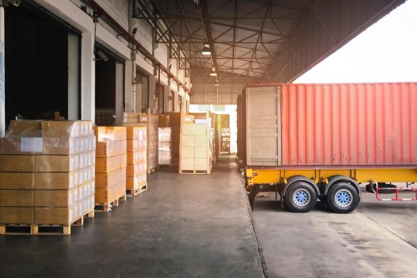 A cargo truck backed in to a loading bay with boxes stacked next to it.