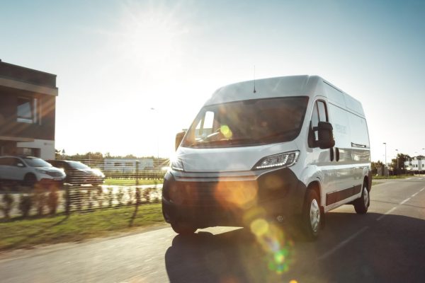 A white commercial van drives down a sunny road next to a business.